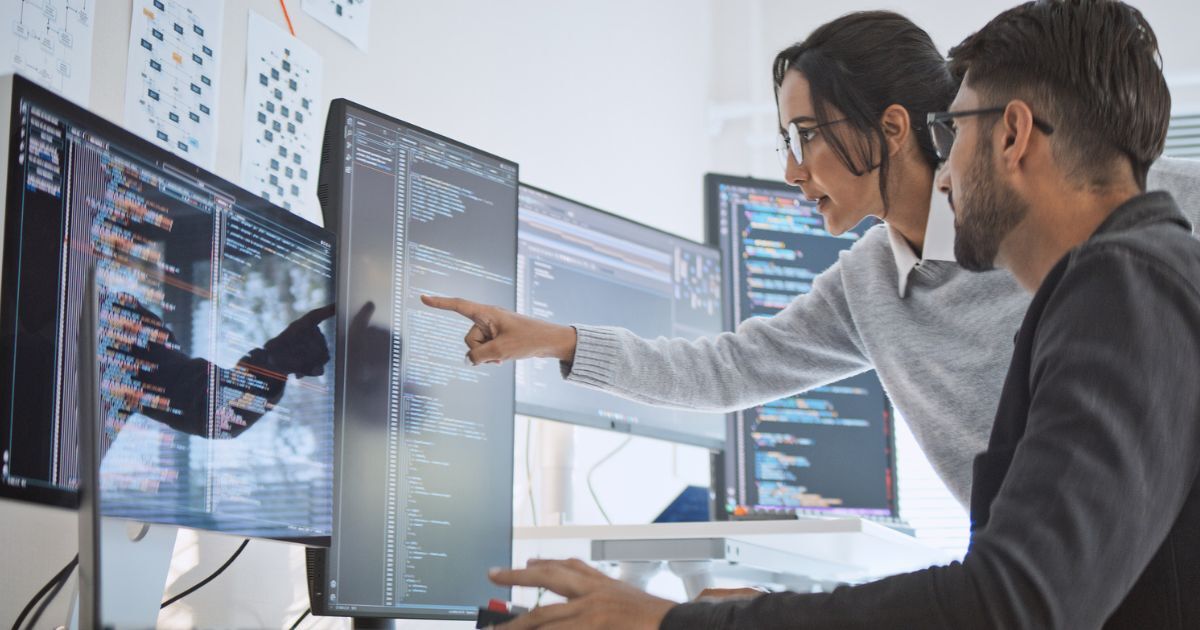 Office setting with several screens on a desk and two colleagues watching and pointing on screen discussing something
