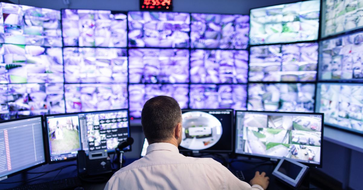 Man in white shirt sits by a desk with a dousin screens in front of him monitoring security cameras